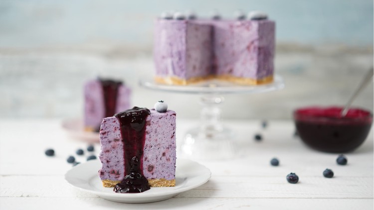 Ein Stück Blaubeer-Torte mit Fruchtsoße auf einem Teller, im Hintergrund die Torte auf einer Glasplatte und eine Schale Soße.