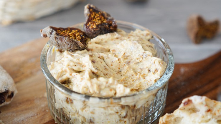 Eine kleine Glasschale mit Feigenbutter, garniert mit getrockneten Feigen, steht auf einem Holzbrett mit geschnittenem Brot.