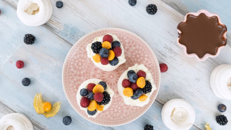 Drei kleine Baiser-Törtchen mit Sahne und Beeren auf einem rosa Teller, umgeben von Obst und unbefüllten Baiserschalen.