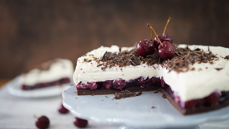 Angeschnittener Kuchen mit Schokoladenboden, Kirschfüllung, Sahnecreme, Schokoraspeln und Kirschen auf einer weißen Tortenplatte.