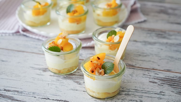 Mehrere kleine Dessertgläser mit Schichten aus Creme, gelben Fruchtstücken, Minzblättern und einer Keksbasis auf Holztisch.