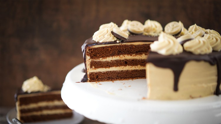 Schokoladentorte mit heller Cremefüllung, Schokoladenglasur, Sahnetuffs und Keksen auf einer weißen Kuchenplatte.