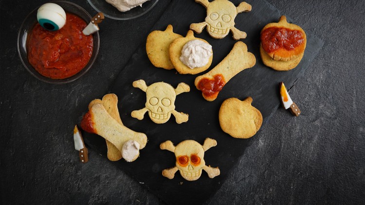 Halloween-Kekse in Form von Kürbissen, Knochen und Totenköpfen auf Schieferplatte, dazu Dips und Messer-Dekoration.