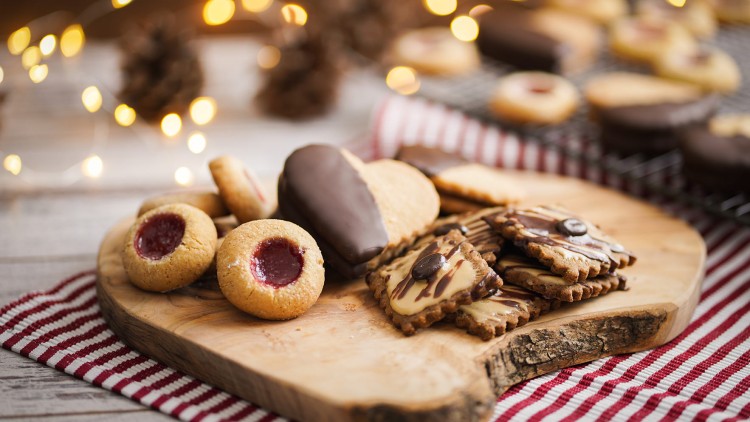 Verschiedene Plätzchen mit Schokolade und Marmelade auf einem Holzbrett vor Lichterkette und Tannenzapfen dekoriert.