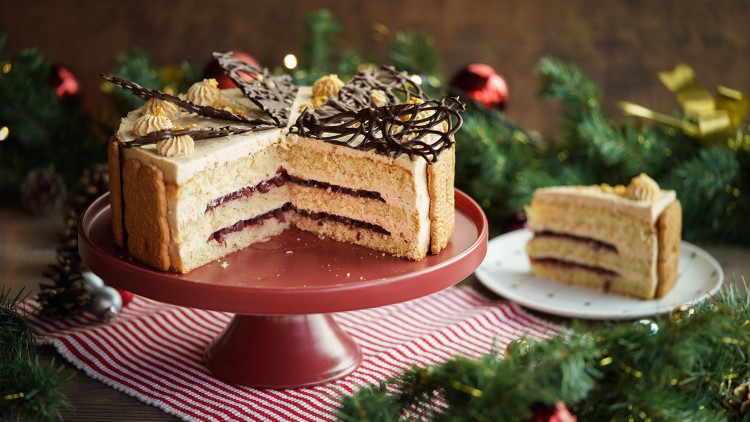 Eine Schichttorte mit Sahne, Marmelade, Schokogitter-Dekor und Keksen auf einer roten Tortenplatte, umgeben von Weihnachtsdeko.