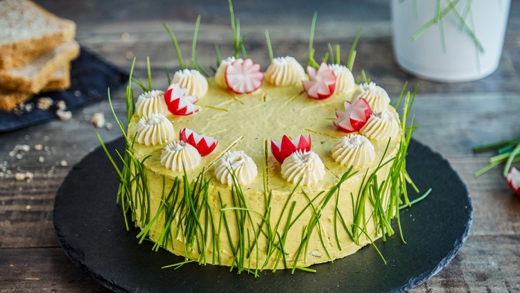 Herzhafte Torte mit gelber Creme, dekoriert mit Schnittlauch, Radieschenblüten und Sahnetuffs auf einer schwarzen Schieferplatte.