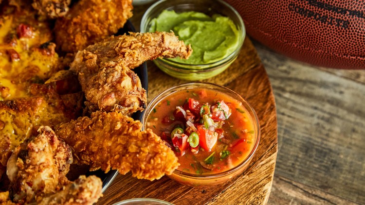 Knusprige Chicken Wings und Nachos mit Schmelzkäse auf einem Holztablett, dazu Guacamole, Salsa und Dip in kleinen Schalen.