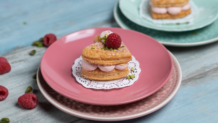 Herzförmiges Blätterteiggebäck mit rosa Creme und Himbeere, dekoriert mit Pistazien, auf rosa und grünen Tellern.