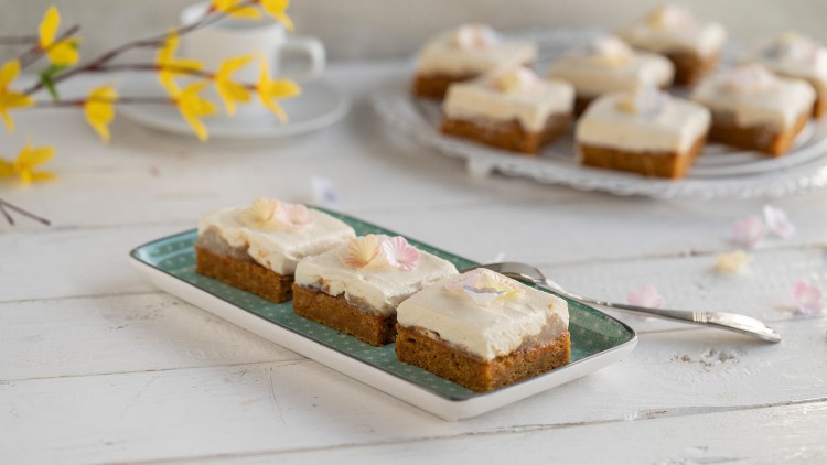 Karottenkuchenstücke mit weißer Creme und essbaren Blüten auf einem rechteckigen Teller, daneben eine Gabel, im Hintergrund Dekoration.