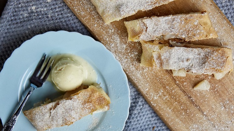 Apfelstrudel auf Holzbrett, geschnittene Stücke mit Puderzucker, Teller mit Apfelstrudel, Kugel Eis, Gabel und Tortenheber.