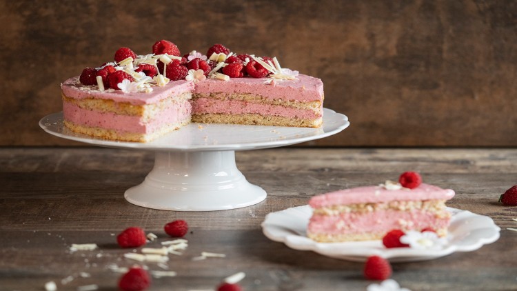 Eine angeschnittene Torte mit rosa Creme, Himbeeren, weißen Schokostreifen und Zuckerblüten auf weißer Tortenplatte.