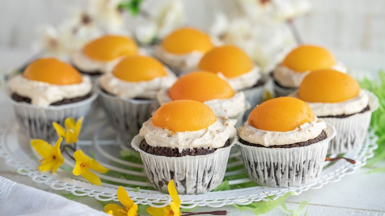 Mehrere Schokoladenmuffins mit Sahnehaube und einer halben Aprikose darauf stehen auf einem weißen Kuchengitter.