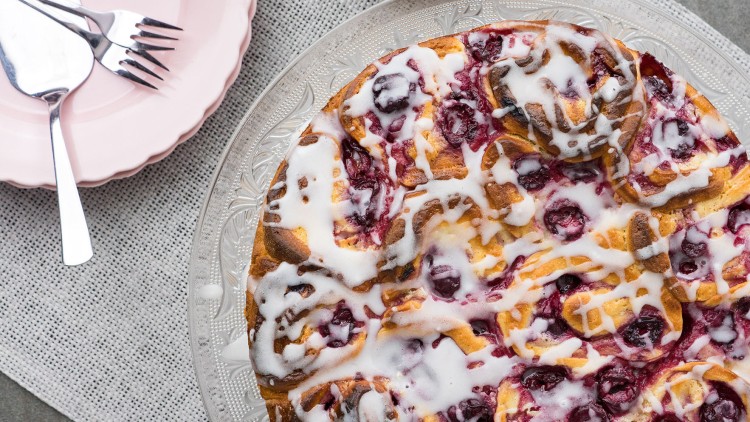 Runder Hefekuchen mit Zuckerglasur und Kirschen auf Glasplatte, daneben rosa Teller mit Besteck auf grauem Stoffuntergrund.