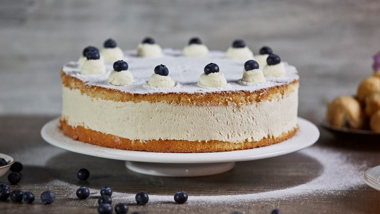 Runde Sahnetorte mit Puderzucker bestäubt, verziert mit Sahnehäubchen und Blaubeeren, auf einem weißen Teller.