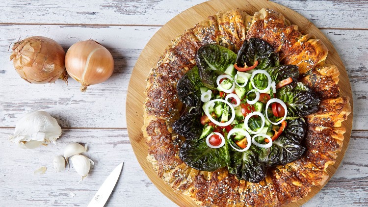 Runder Holz-Teller mit Salat aus Gurken, Tomaten, Zwiebeln und Blattsalat, umringt von gefülltem, goldbraunem Gebäck.