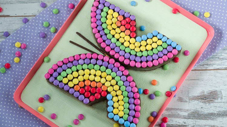 Ein Schmetterlingskuchen aus zwei Stücken, verziert mit bunten Schokolinsen auf einem Tablett mit bunten Streudekorationen.