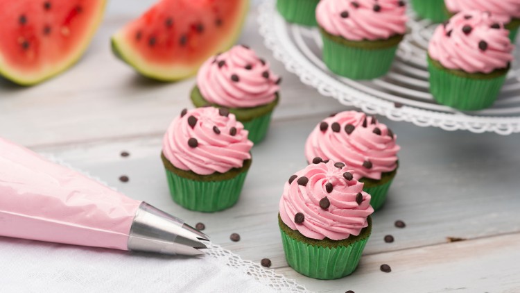 Mehrere grüne Cupcakes mit rosa Creme, Schokotropfen und einer Tortenspritze auf Holzuntergrund, im Hintergrund Wassermelonenscheiben.