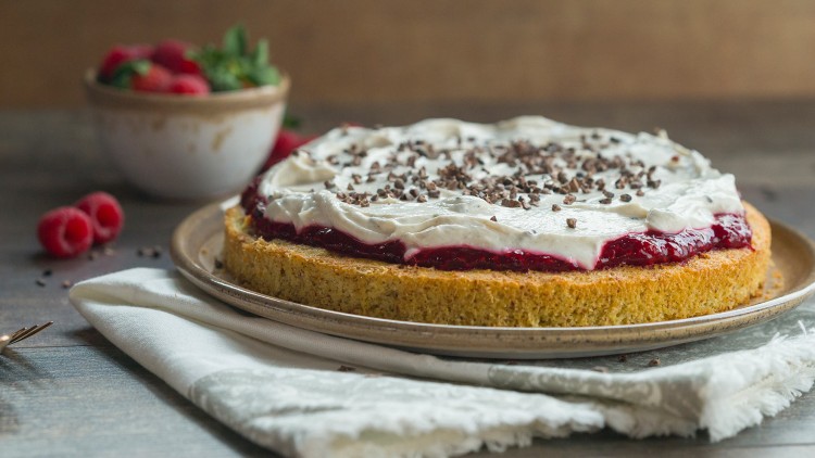 Runder Kuchen mit Schicht aus roter Creme, weißer Schlagsahne und Schokoraspeln auf einem Teller, daneben Himbeeren in einer Schale.