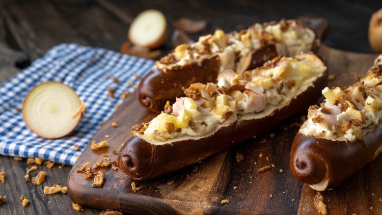 Gefüllte Laugenstangen mit cremigem Belag, Apfelwürfeln und gerösteten Zwiebeln auf einem Holzbrett, daneben eine halbe Zwiebel.
