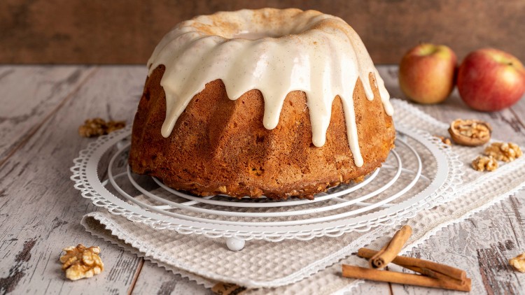 Gugelhupfkuchen mit hellem Zuckerguss auf einem weißen Gitter, dekoriert mit Walnüssen, Äpfeln und Zimtstangen.