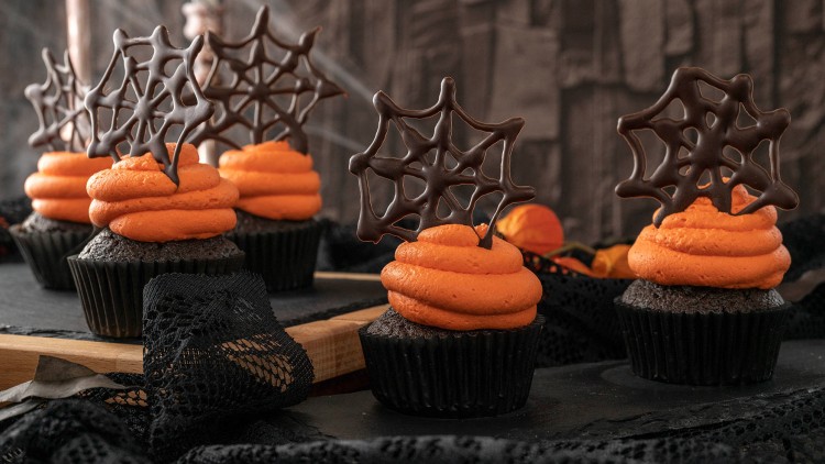 Schokoladen-Cupcakes mit orangefarbener Cremehaube und Schoko-Dekor in Spinnennetzform auf schwarzem Untergrund.