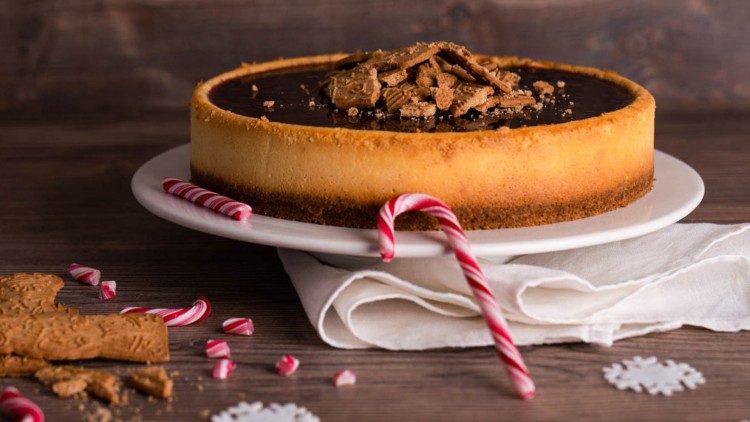 Runder Käsekuchen mit Schokoglasur und Keksstücken, dekoriert mit Zuckerstangen auf einem weißen Teller und Holzuntergrund.
