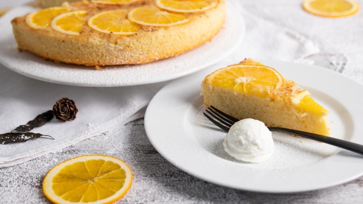 Ein gedeckter Tisch mit Orangenkuchen auf einer Tortenplatte, ein Kuchenstück mit Sahnekugel auf einem weißen Teller.