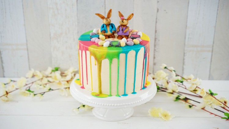 Bunte Drip-Torte mit Schoko-Osterhasen-Deko, Mini-Eiern und Blumen auf weißer Tortenplatte vor hellem Holz-Hintergrund.