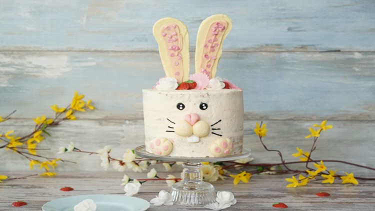 Eine Torte mit Hasengesicht, Schokohasenohren, rosa Dekoration und Erdbeeren auf einem Glasständer vor hellblauem Hintergrund.