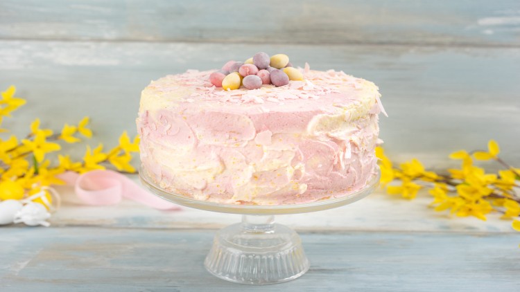 Runde Torte mit rosa-gelber Creme, Schokoladeneiern und Streuseln, auf Glasplatte mit gelben Blüten dekoriert.