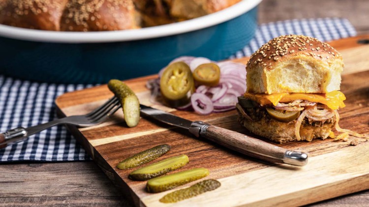 Ein Burgerbrötchen mit Sesam, Hackfleisch, Käse, Zwiebeln und Jalapeños auf Holzbrett mit Gewürzgurken und Besteck.