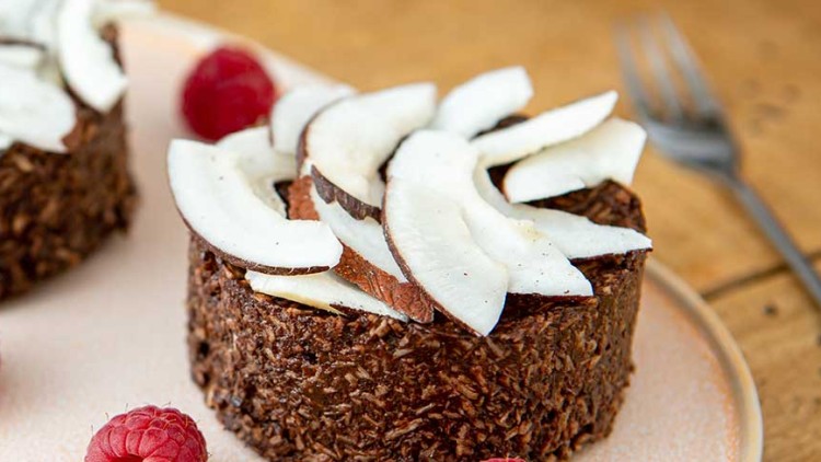 Zwei Schoko-Kokostörtchen mit frischen Kokoschips und Himbeeren auf einem Teller, daneben eine Gabel auf Holztisch.