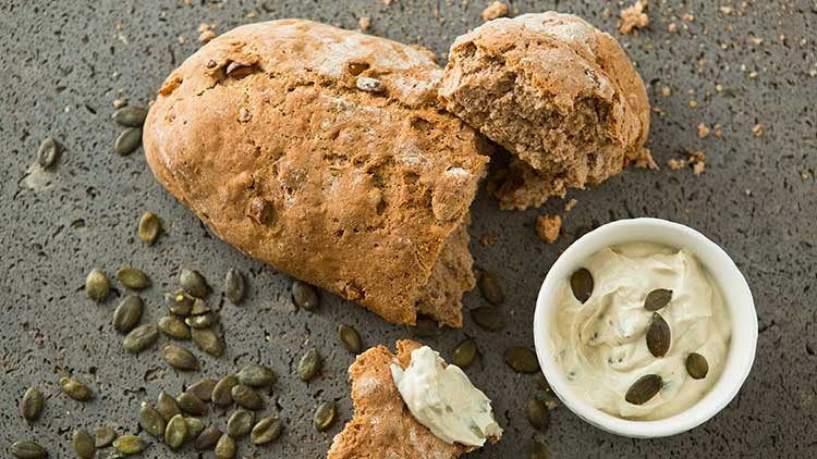 Ein aufgebrochenes Vollkornbrötchen mit Kürbiskernen, daneben eine Schale mit cremigem Aufstrich und Kürbiskernen auf grauem Untergrund.