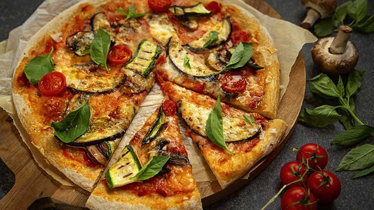 Eine vegetarische Pizza mit Tomatensauce, Zucchini, Champignons, Basilikum und Kirschtomaten auf einem Holzbrett.
