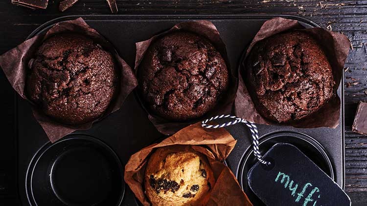 Drei Schokoladenmuffins in einem Backblech und ein heller Muffin in Papier mit angehängtem Schild, auf dem „muffin“ steht.