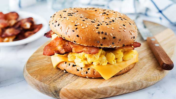 Ein Sesam-Bagel belegt mit Rührei, einer Scheibe Käse und knusprigem Bacon liegt auf einem runden Holzbrett.