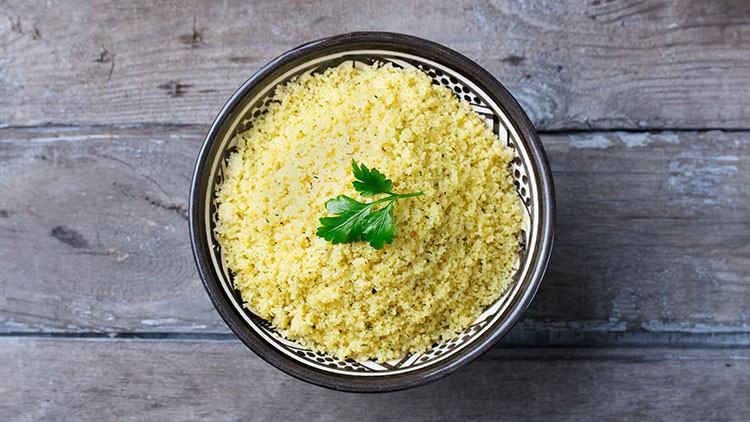 Eine Schale mit gelbem Couscous und einem kleinen Zweig frischer Petersilie steht auf einem grauen Holztisch.