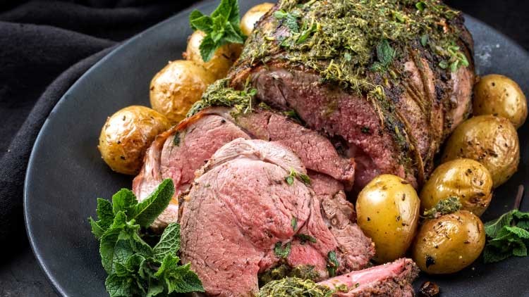 Ein aufgeschnittenes Lammfilet mit Kräutern, umgeben von kleinen goldbraunen Kartoffeln und frischer Minze auf einem schwarzen Teller.