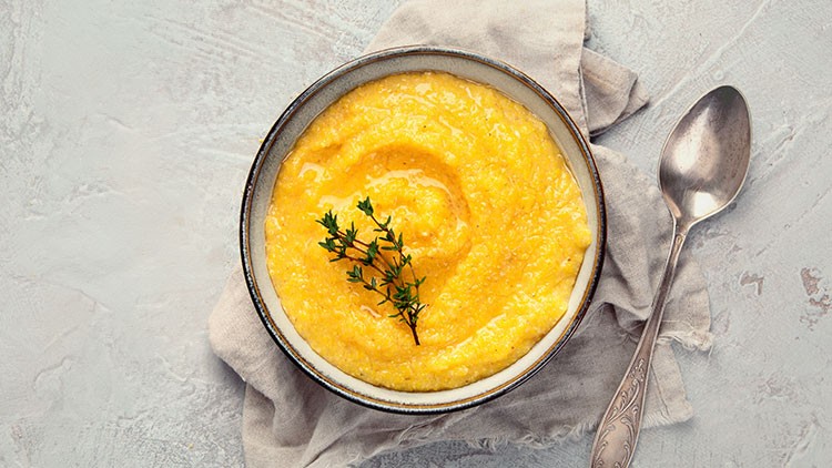 Eine Schale mit gelber Polenta, dekoriert mit frischem Thymian, steht auf einem beigen Tuch neben einem silbernen Löffel.