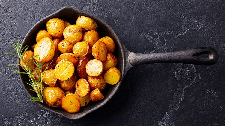 Gebratene Kartoffelscheiben und Rosmarinzweig in einer schwarzen Pfanne auf dunklem Untergrund.