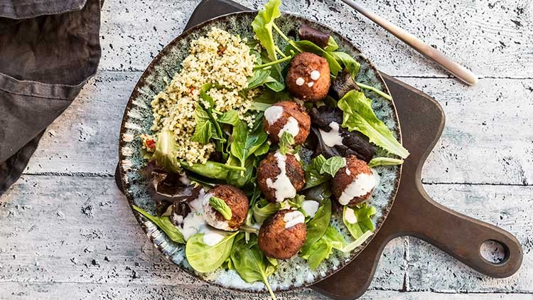 Fünf Falafel-Bällchen mit weißem Dressing liegen auf grünem Salat, daneben Couscous auf einem rustikalen Teller.