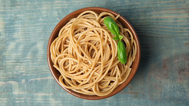 Eine braune Schale mit gekochten Vollkornspaghetti, garniert mit zwei Basilikumblättern, steht auf einer hellblauen Holzoberfläche.