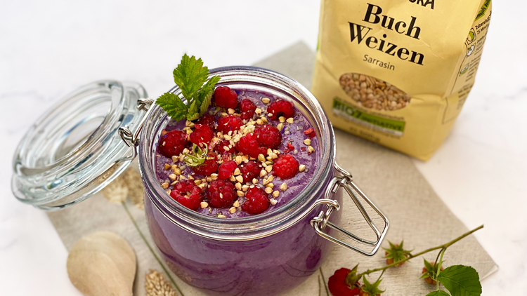 Ein Glas mit lila Smoothie, dekoriert mit Himbeeren, Buchweizen und Minzblatt, steht neben einer Packung Buchweizen.