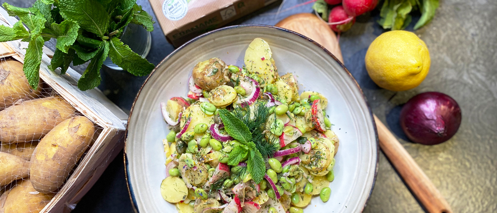 Ein Teller mit Kartoffelsalat, Edamame, Dill, roten Zwiebeln und Minze steht neben einer Zitrone, Kartoffeln und Radieschen.