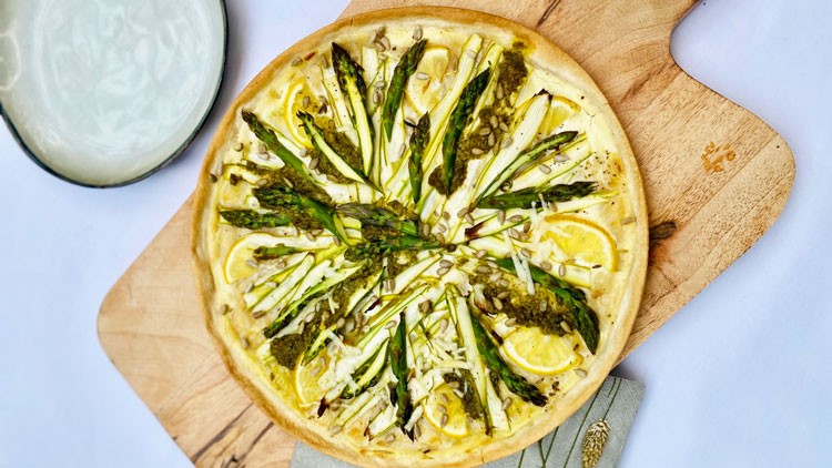 Runde Spargelquiche mit grünen Spargelstangen, Zitronenscheiben und Käse auf einem Holzbrett, daneben ein weißer Teller.