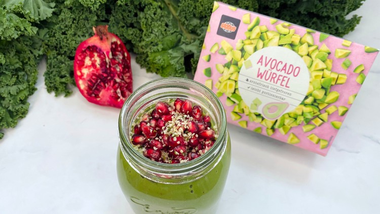 Grüner Smoothie im Glas mit Granatapfelkernen und Samen sowie einem halben Granatapfel und einer GLOBUS Avocado-Würfel-Packung.