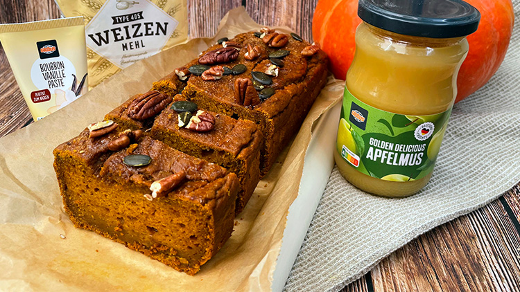 Ein angeschnittener Kürbiskuchen mit Nüssen und Kernen, daneben Apfelmus im Glas und Zutaten wie Mehl und Vanillepaste.