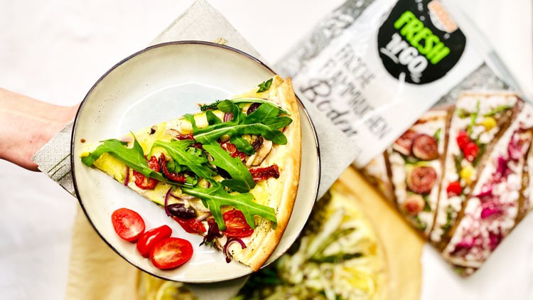 Ein Teller mit einem Stück Gemüsepizza, Rucola und halbierten Tomaten, im Hintergrund die Verpackung von GLOBUS Fresh to Go.