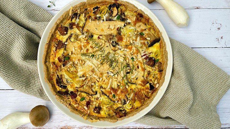 Eine herzhafte Quiche mit Pilzen, Lauch, Rosmarin und Pinienkernen auf hellem Holztisch, daneben ein Stofftuch und Kräuterseitlinge.