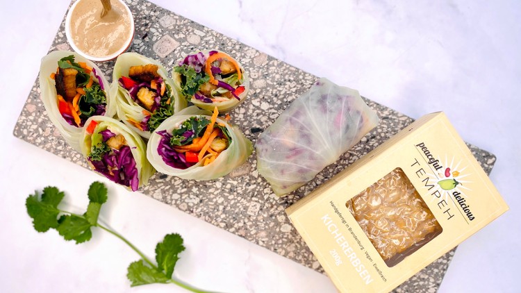 Reispapierrollen mit buntem Gemüse und Tempeh, daneben eine Schale Dip, frischer Koriander und eine Tempeh-Verpackung aus Kichererbsen.